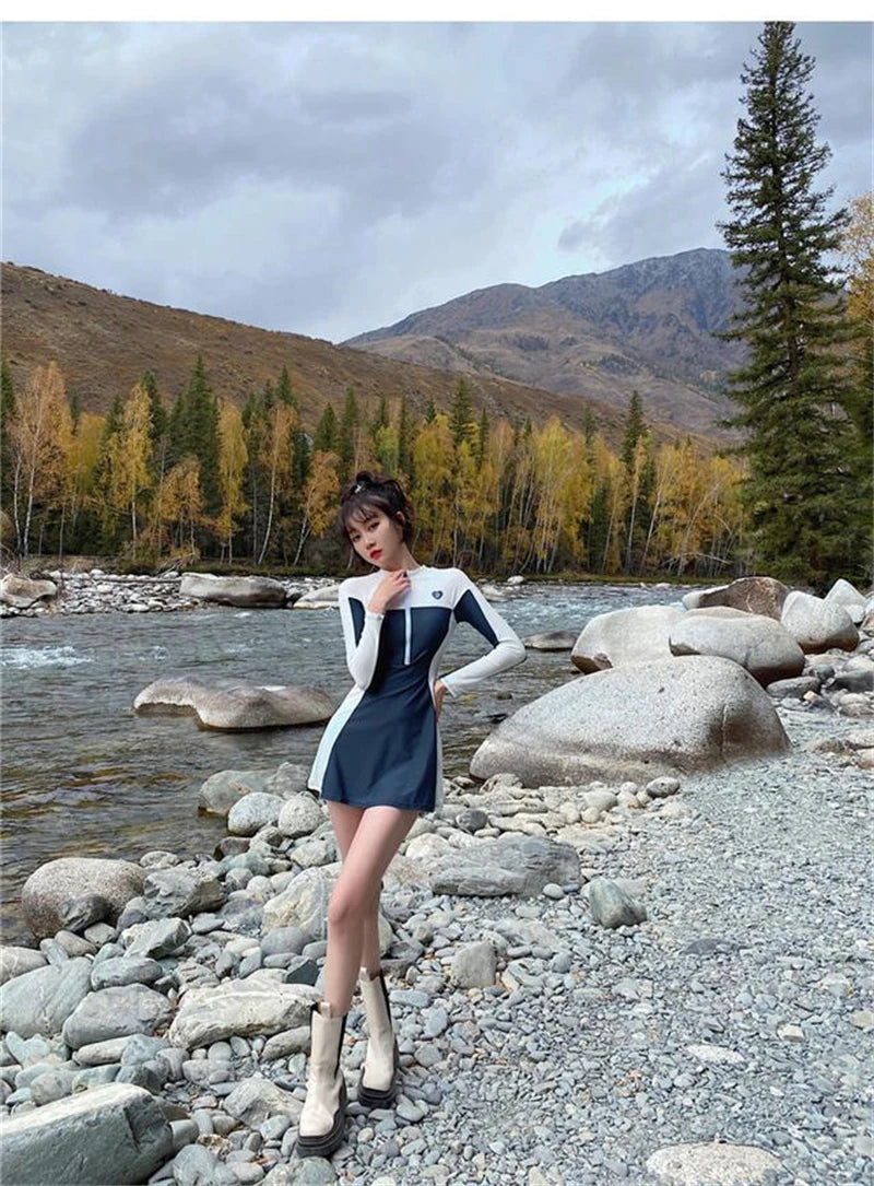 Une jeune femme à la mode posant au bord d'une rivière rocheuse dans un maillot de bain femme sport BREEZEBEEZ unique, avec des arbres aux couleurs d'automne et des montagnes lointaines en arrière-plan.