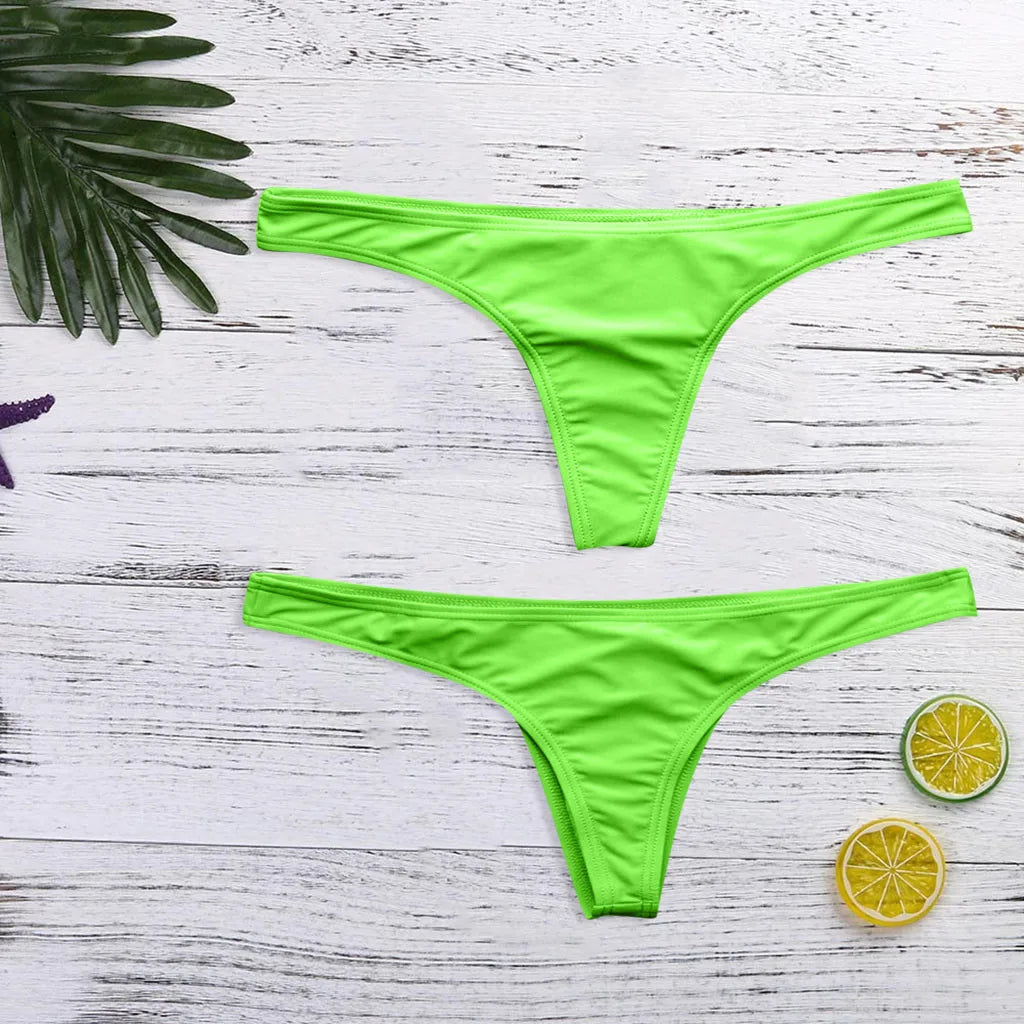 Une paire de Bas Maillot de Bain Femme 1 Pièce vert vif disposés à plat en forme de V inversé sur un fond en bois blanc, flanqué de feuilles tropicales et de citrons tranchés, suggère un thème estival et prêt pour la plage de BREEZE BEEZ.