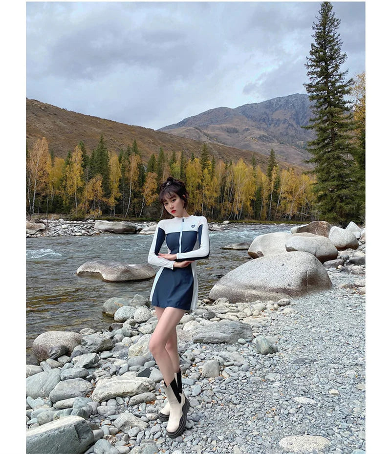 Une femme debout au bord d'une rivière dans un très beau paysage de montagne, regardant pensivement un BREEZEBEEZ Maillot de Bain Femme Sport qu'elle tient.