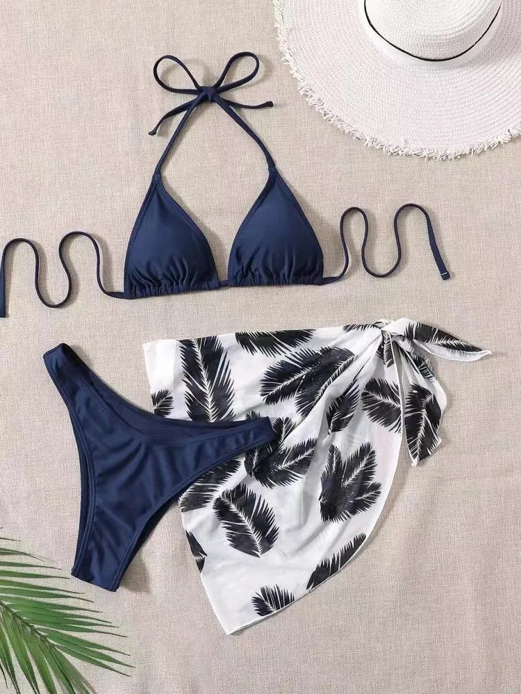 Un très beau Maillot de Bain Femme 3 Pièces | Haut de bikini Ensemble haut & Bas associé à des bas assortis et un paréo à imprimé tropical blanc et noir, soigneusement disposés sur une surface beige aux côtés d'un chapeau de soleil blanc et d'une feuille de palmier verte, transmettant une ambiance BREEZE BEEZ.