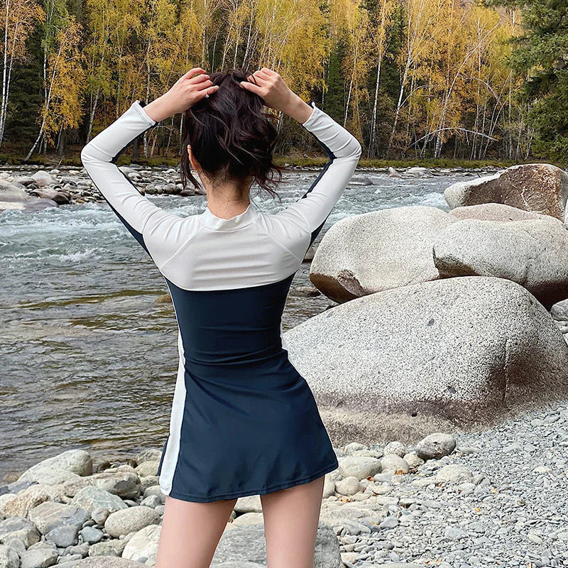 Une personne vêtue d'une tenue blanche et bleu marine se tient au bord d'une rivière pittoresque, entourée de gros rochers et d'arbres automnaux, attachant ses cheveux tout en contemplant la nature et profitant des vacances avec son BREEZEBEEZ Maillot de Bain Femme Sport.