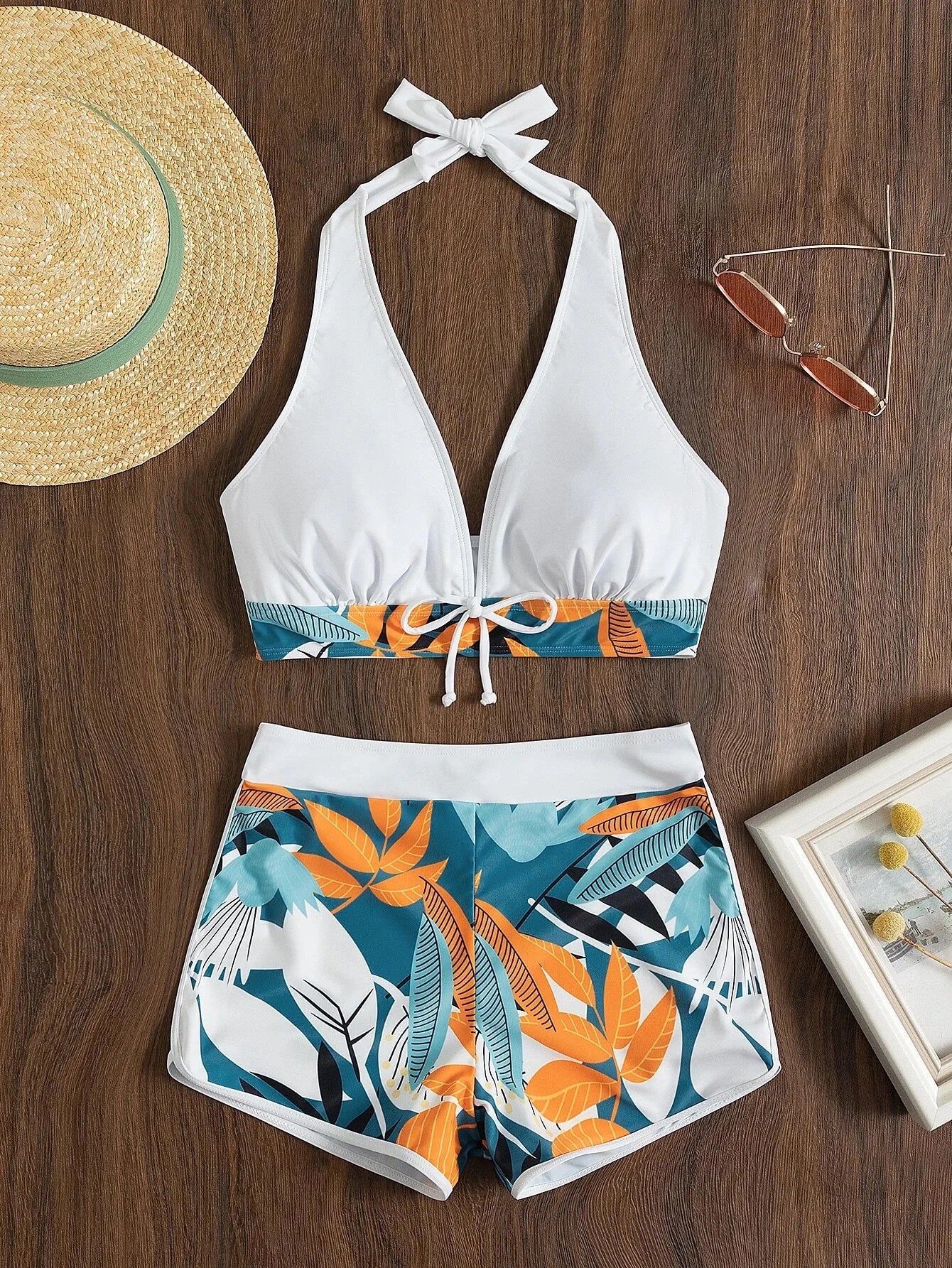 Une présentation à plat d'une très belle tenue d'été comprenant un bikini haut dos nu blanc avec des détails rayés et un devant noué de BREEZE BEEZ, associé à un short Maillot de Bain Short Femme 2 pièces sexy à imprimé tropical, complété par des accessoires dont un chapeau de paille.