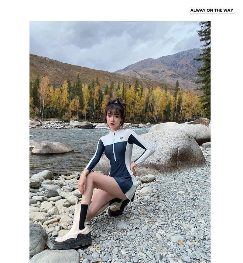 Une personne élégante posant sur des rochers de rivière sur fond d'arbres automnaux et d'un paysage de montagne nuageux, portant un BREEZEBEEZ Maillot de Bain Femme Sport unique.